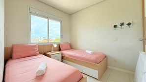 A bedroom with two beds, a window, a bedside table, and a wall-mounted light fixture.