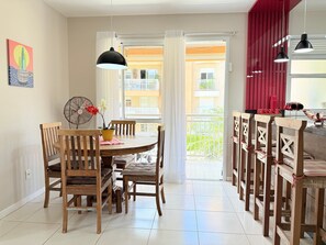A dining area with a round table, wooden chairs, a fan, and a view of a balcony with red curtains. - Residencial com piscina (Florianópolis)