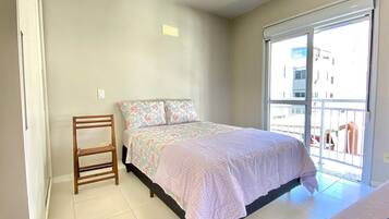 Un dormitorio con cama, una silla de madera, una ventana con vista a edificios y una puerta.