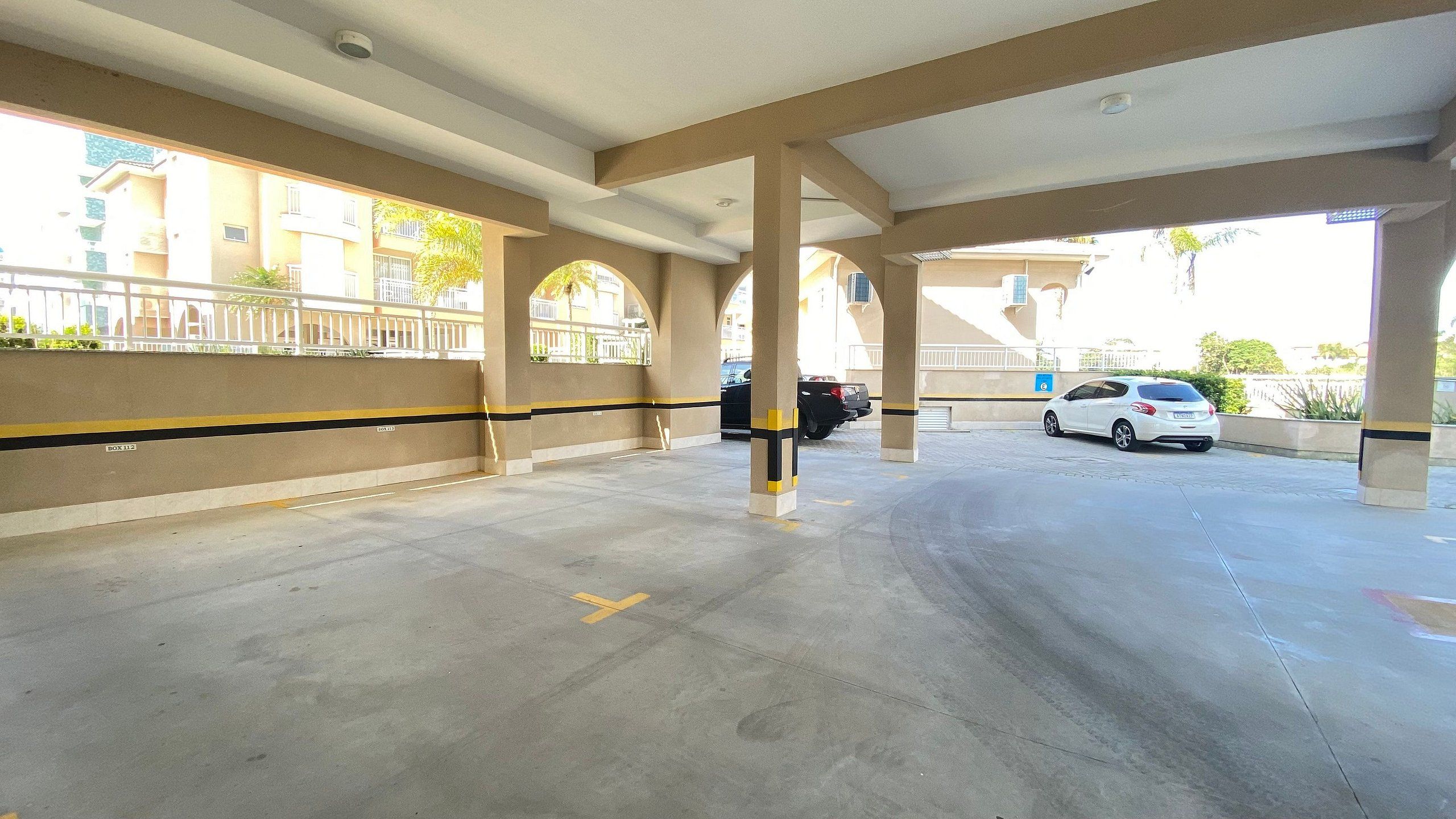 A covered parking area with concrete floors and pillars, a white car parked, and a building with balconies in the background.