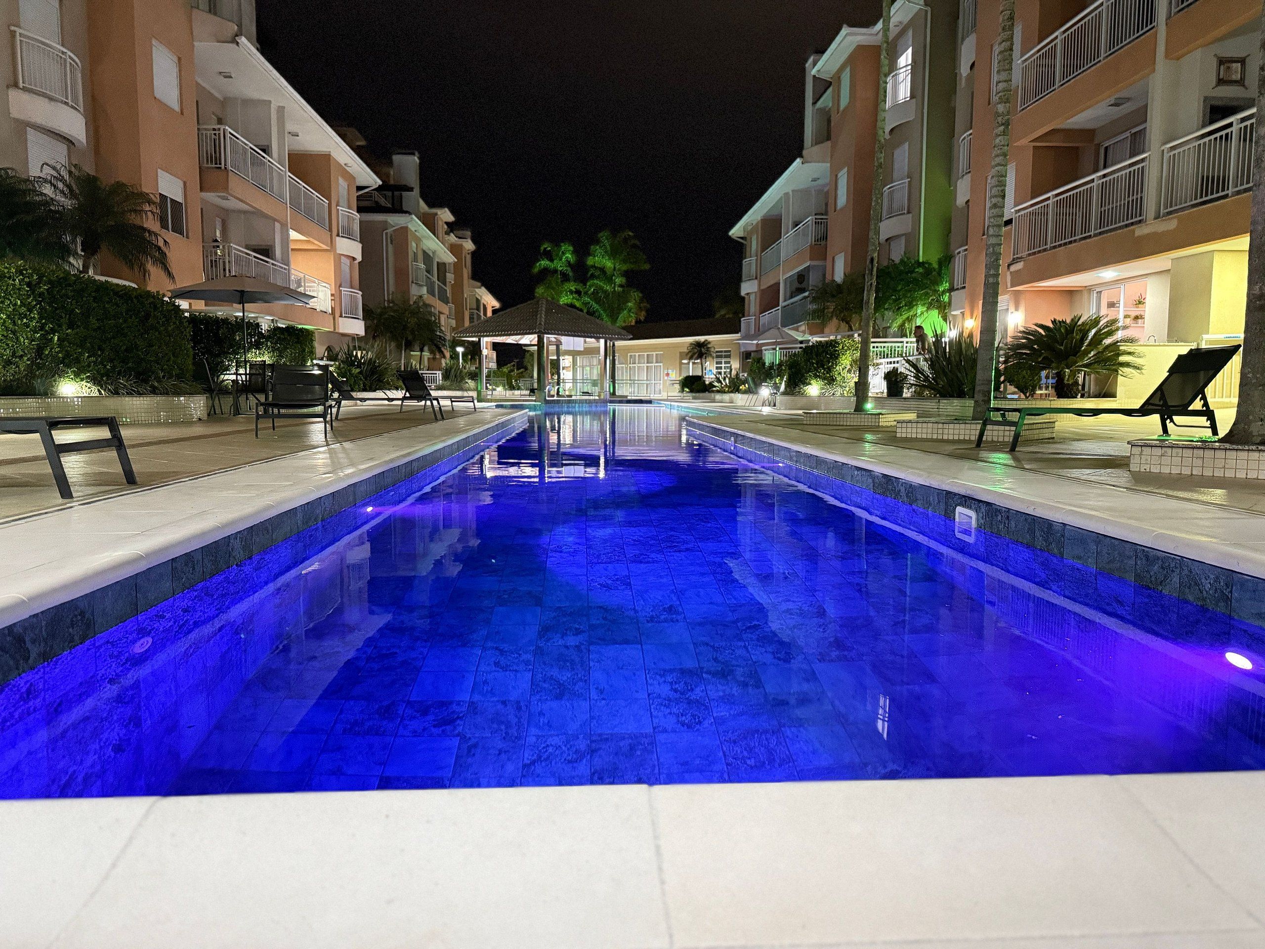 A swimming pool with a blue light, surrounded by buildings with balconies and outdoor seating.