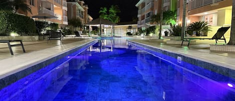 A swimming pool with a blue light, surrounded by buildings with balconies and outdoor seating.