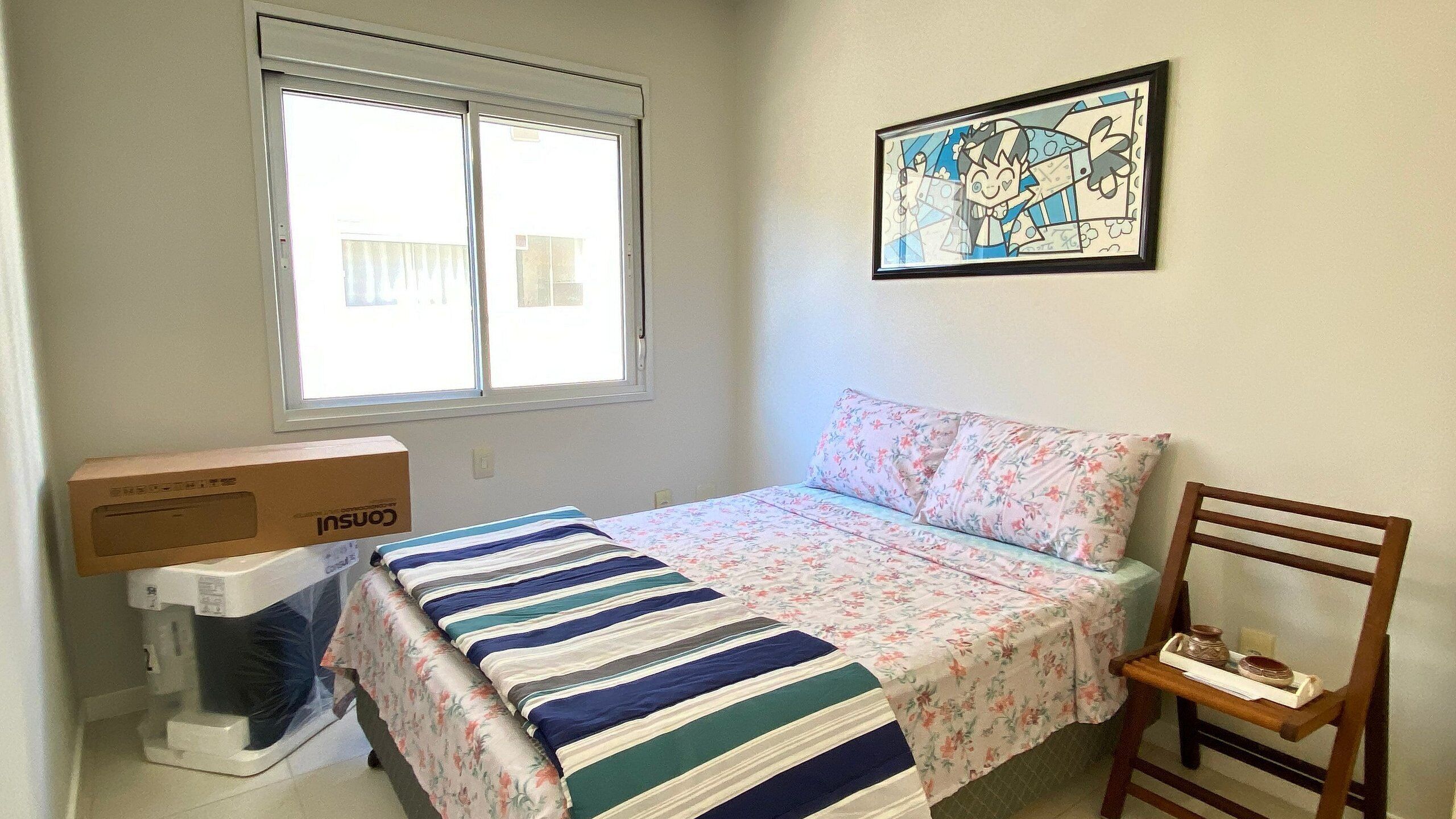 A bedroom with a bed, a window, a framed picture, a chair, and a box.