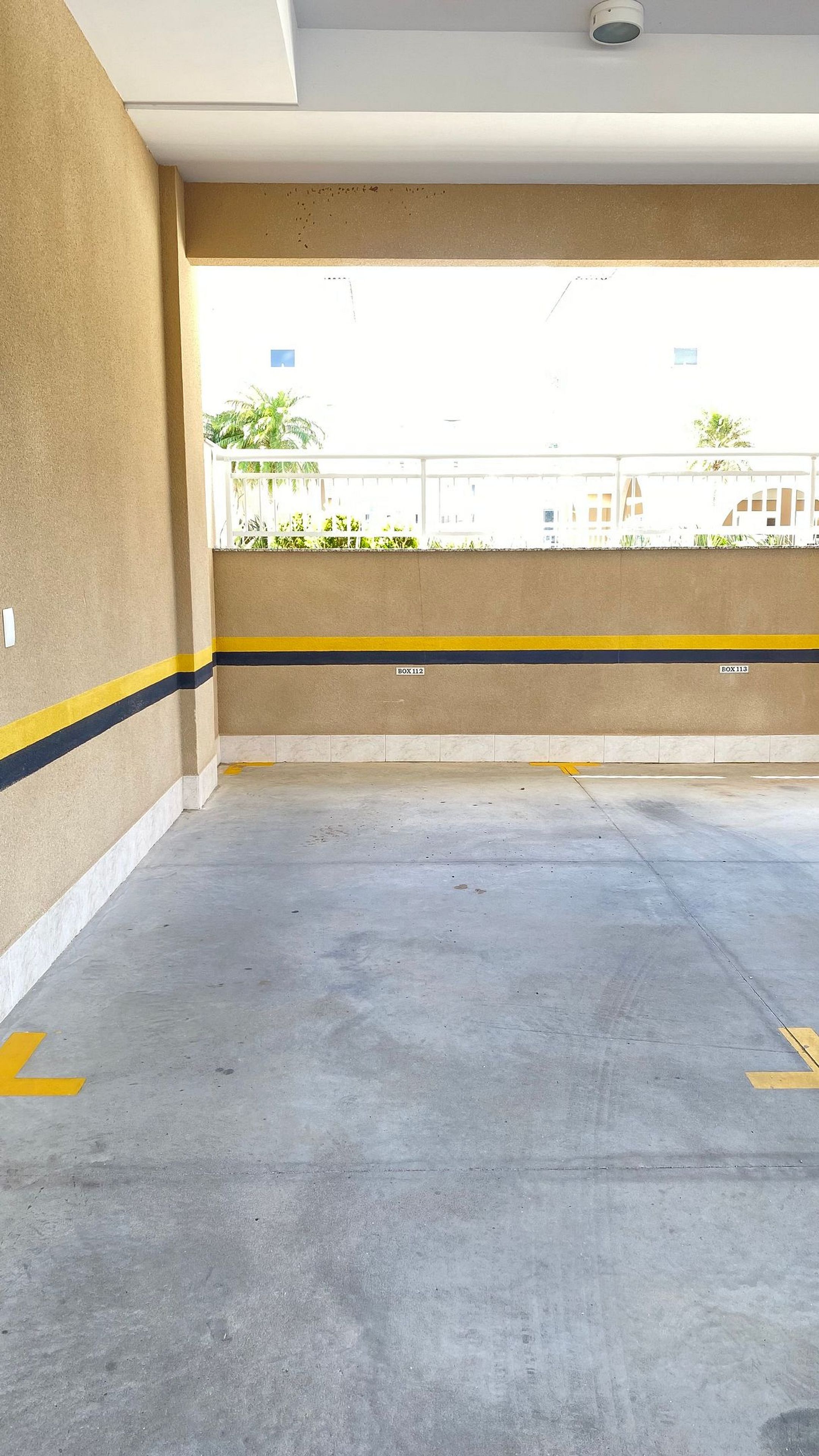 Un estacionamiento vacío con pisos y paredes de concreto, espacios de estacionamiento delimitados y vistas a edificios y áreas verdes en el exterior.