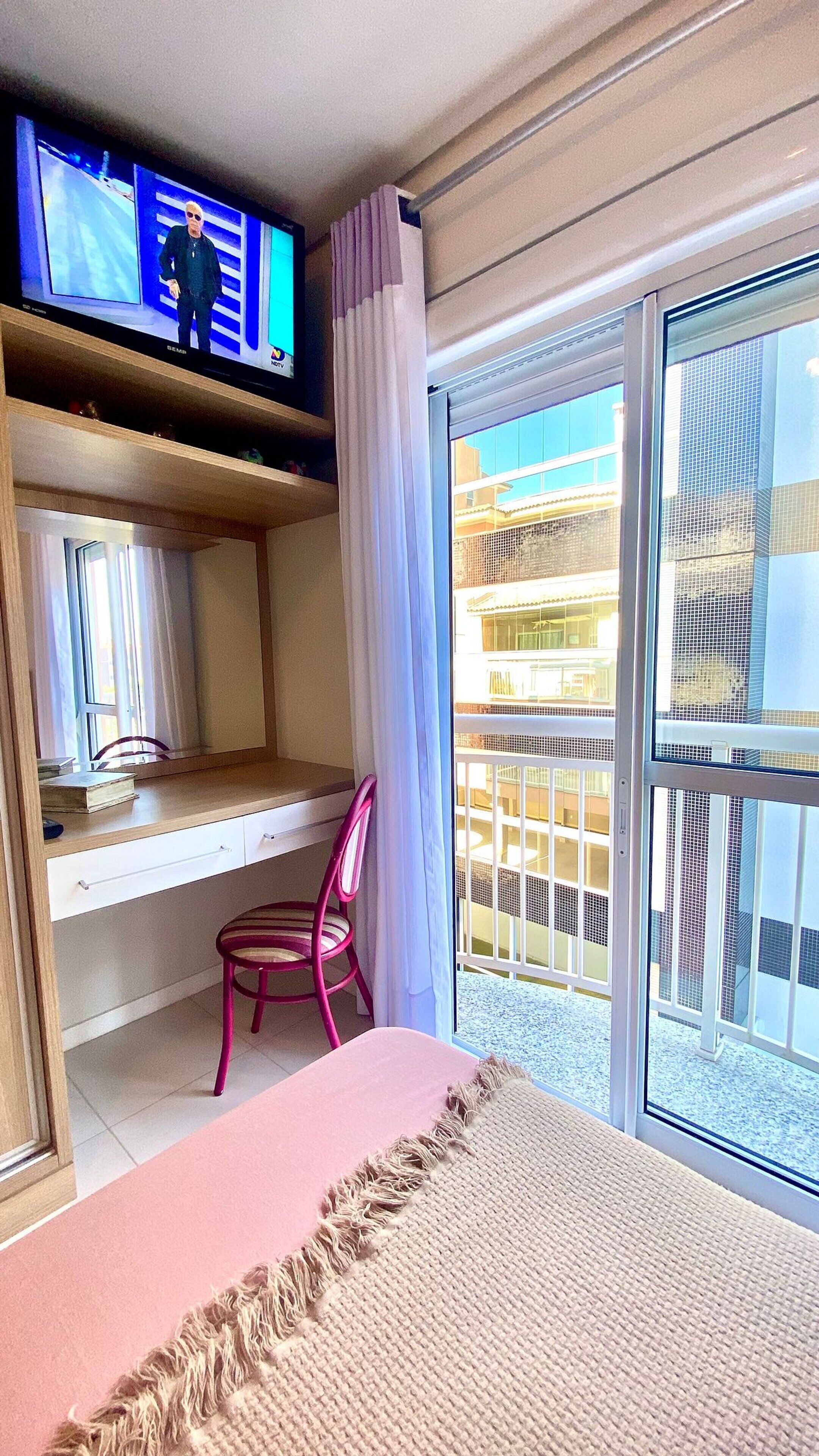 A room with a bed, a TV on a shelf, a chair, and a balcony with a view of buildings.