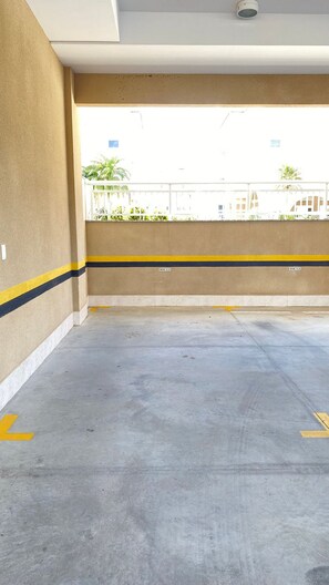 An empty parking garage with concrete floors and walls, marked parking spaces, and a view of buildings and greenery outside.