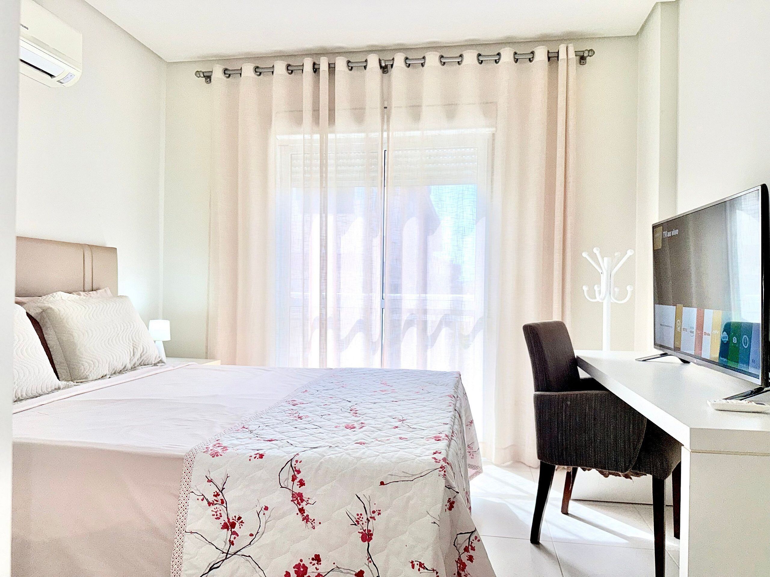 A hotel room with a bed, a desk with a TV, and a window with curtains.