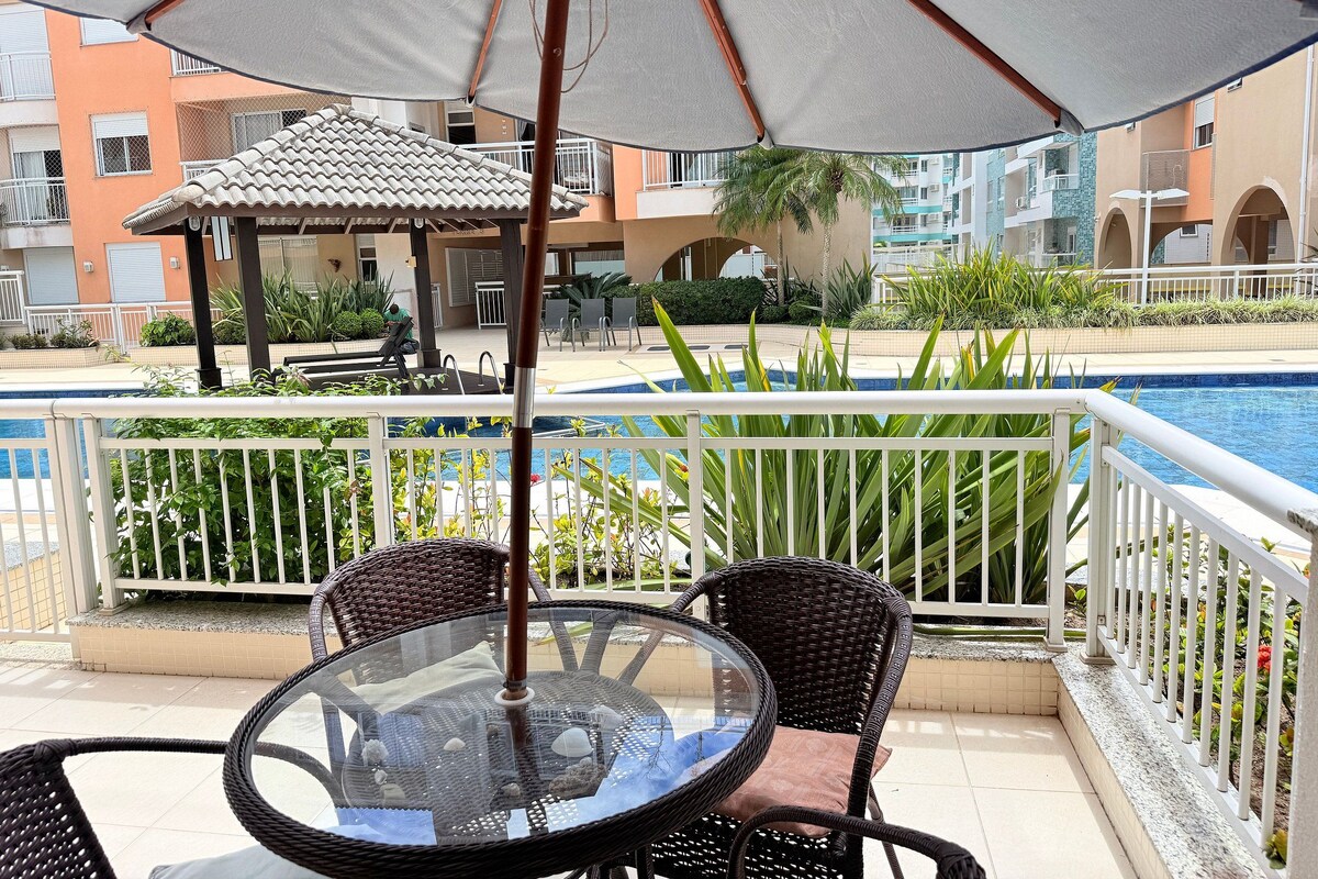 A glass table with wicker chairs, an umbrella, and a poolside view of a building with balconies.