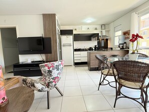A modern kitchen with a flat-screen TV, a refrigerator, and a dining area with wicker chairs and a glass table. - Residencial com piscina (Florianópolis)