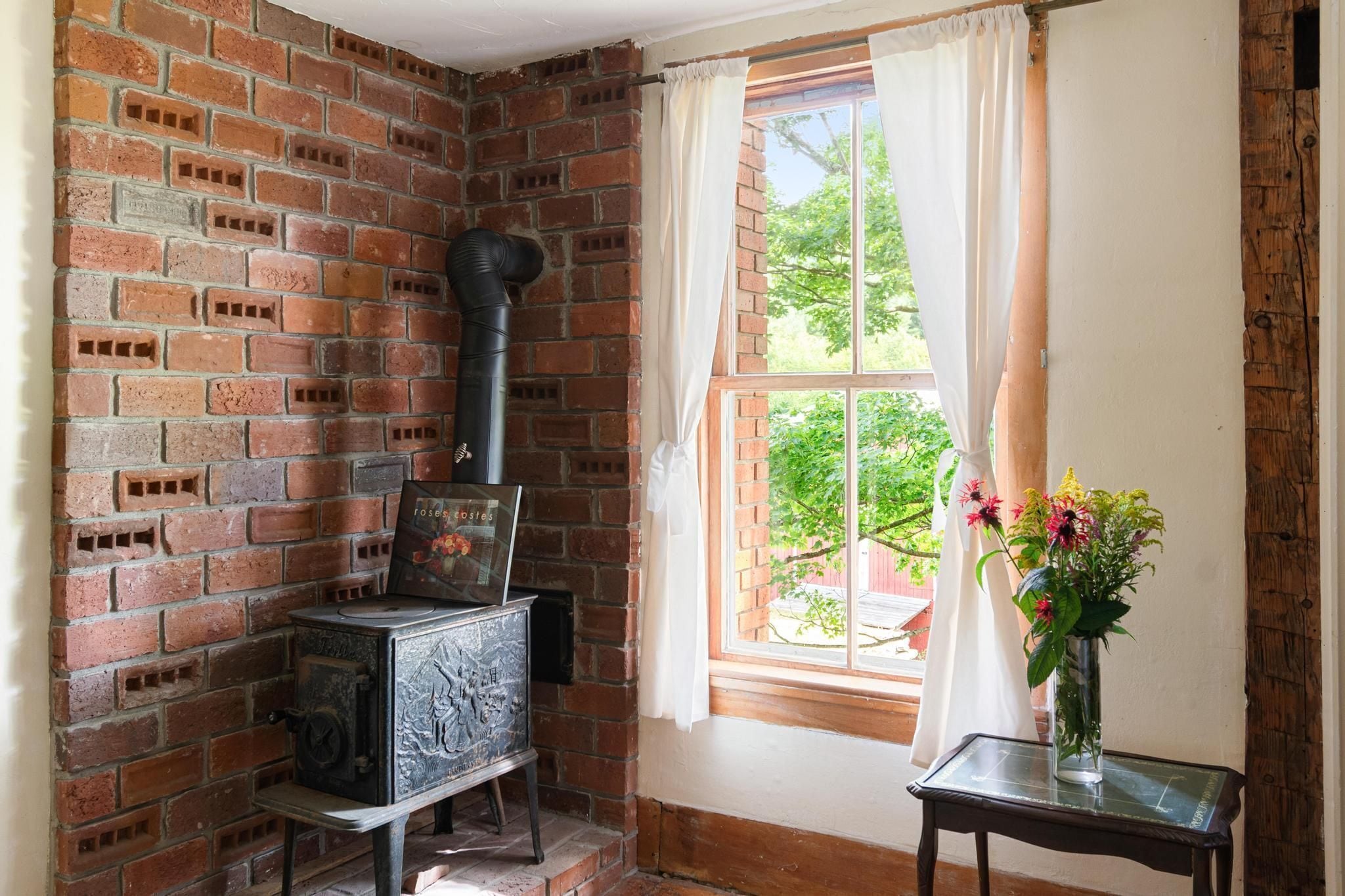 A room with a brick wall, a wood-burning stove, a window with curtains, and a vase with flowers on a table.