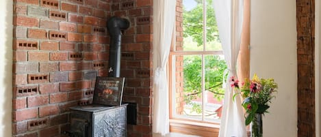 A room with a brick wall, a wood-burning stove, a window with curtains, and a vase with flowers on a table.