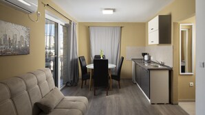 Ett kompakt bostadsutrymme med en soffa, ett matbord, stolar, en liten köksdel och ett stort fönster med gardiner.