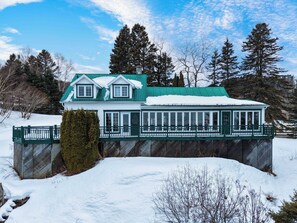 A two-story house with a green roof and white walls, surrounded by snow and trees.