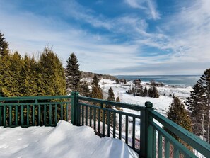 Un paysage enneigé avec une terrasse en bois et une vue sur un lac et des bùtiments au loin.