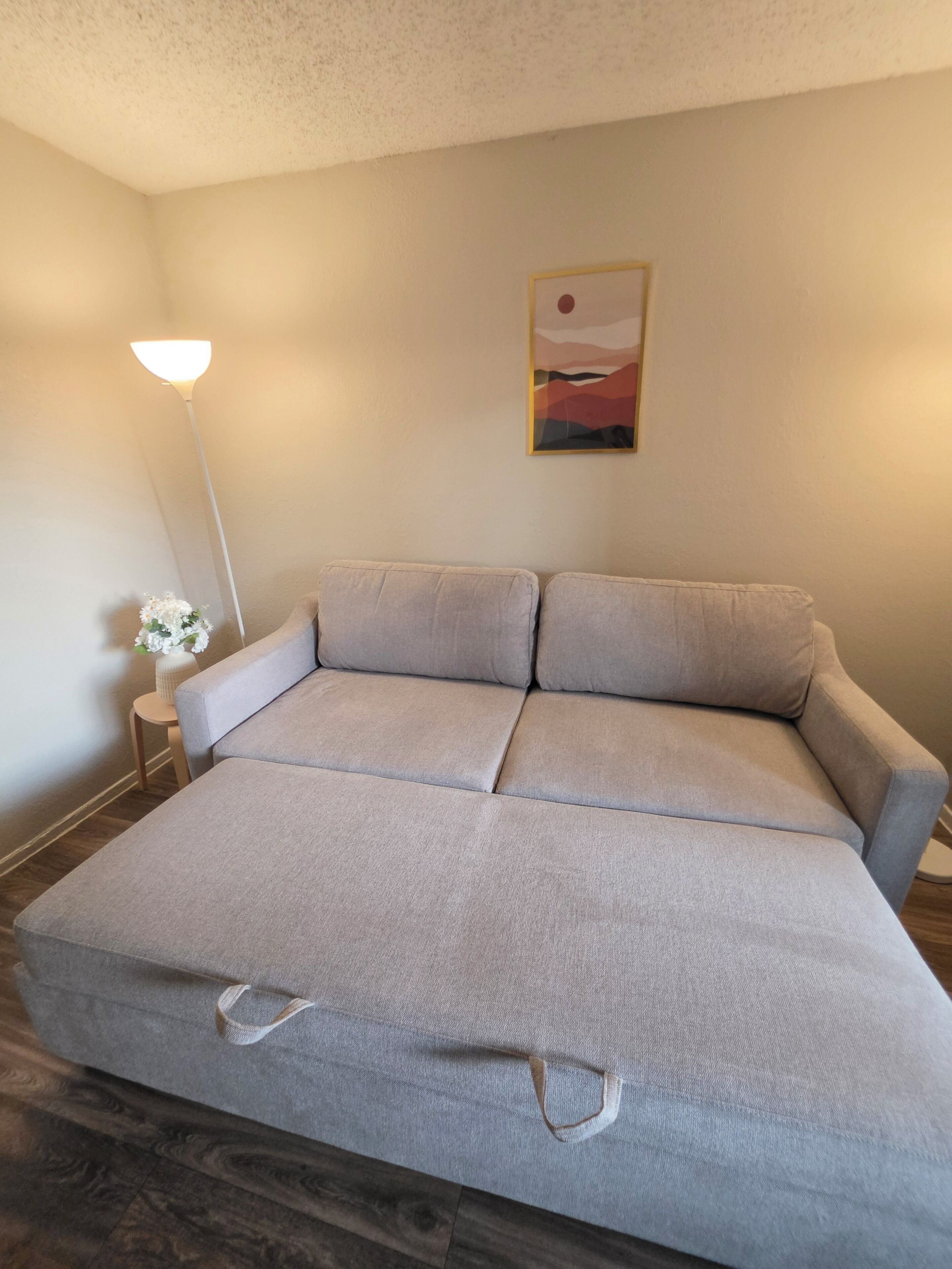 A room with a large sofa, a small side table with a vase of flowers, a floor lamp, and a painting on the wall.