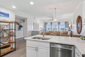 A modern kitchen with a central island, a dining area with wicker furniture, and a view of the outdoors. - Spacious beachfront condo with gulf views (Miramar Beach)