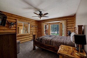 A log cabin bedroom with a bed, a ceiling fan, a wooden desk, and a window with a view of trees.