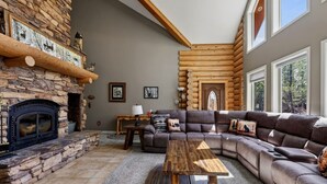 A log cabin interior with a stone fireplace, a large sectional sofa, and a wooden table. - Golf Course Cabin | Sleeps 16, Hot Tub, Game Room, Golf, 25min to YSNP (Island Park)
