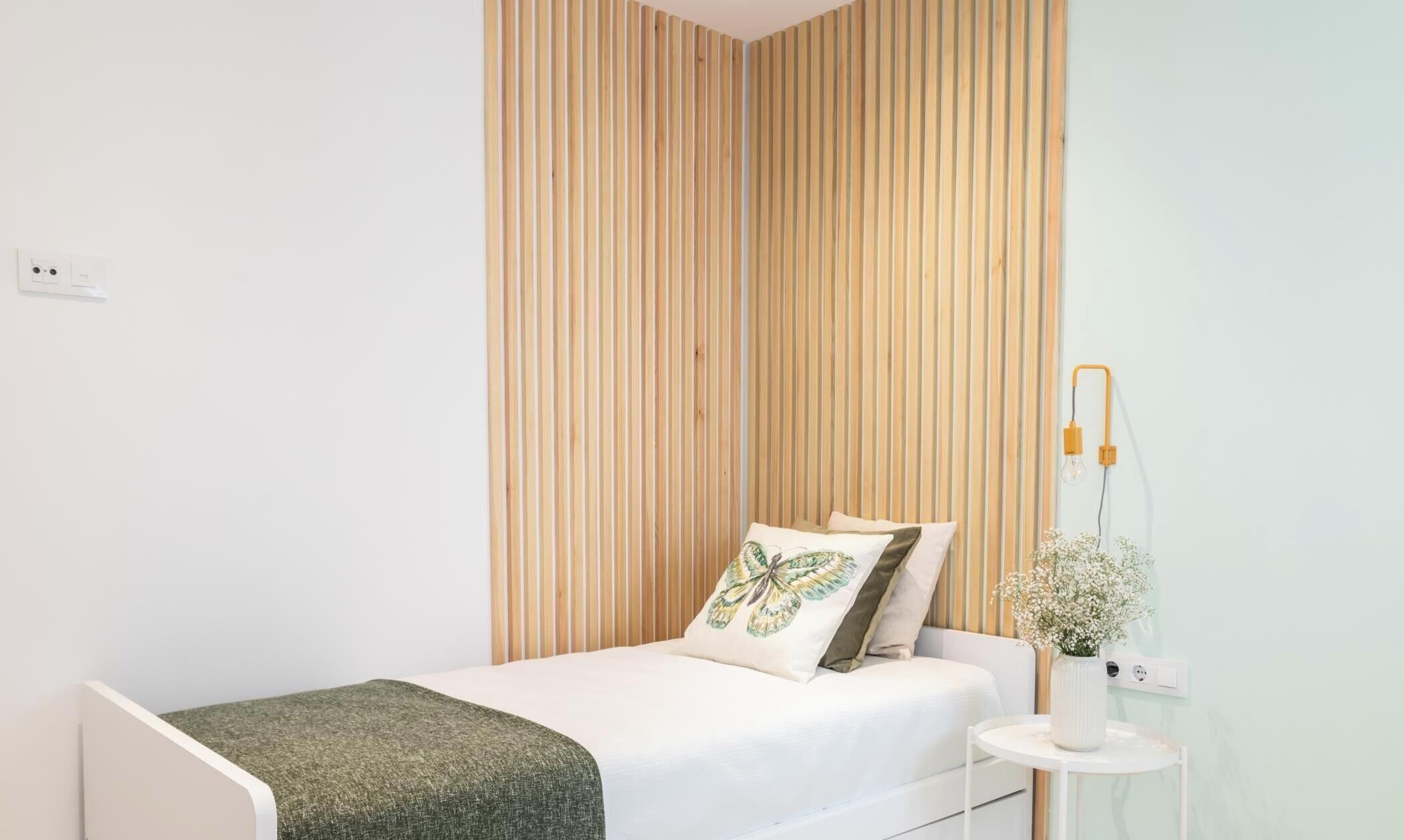 A single bed with a green throw blanket, a wooden slat wall, a bedside table with a vase of flowers, and a wall-mounted lamp.