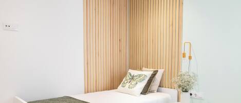 A single bed with a green throw blanket, a wooden slat wall, a bedside table with a vase of flowers, and a wall-mounted lamp.