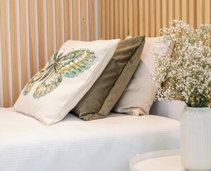 Ein Bett mit weißer Matratze, zwei Kissen, einer Vase mit weißen Blumen und einer Holzhintergrundwand.