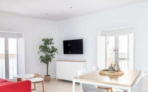 Ein helles, modernes Wohnzimmer mit Essbereich, Fernseher, Topfpflanze und Balkon, der durch die Tür sichtbar ist.