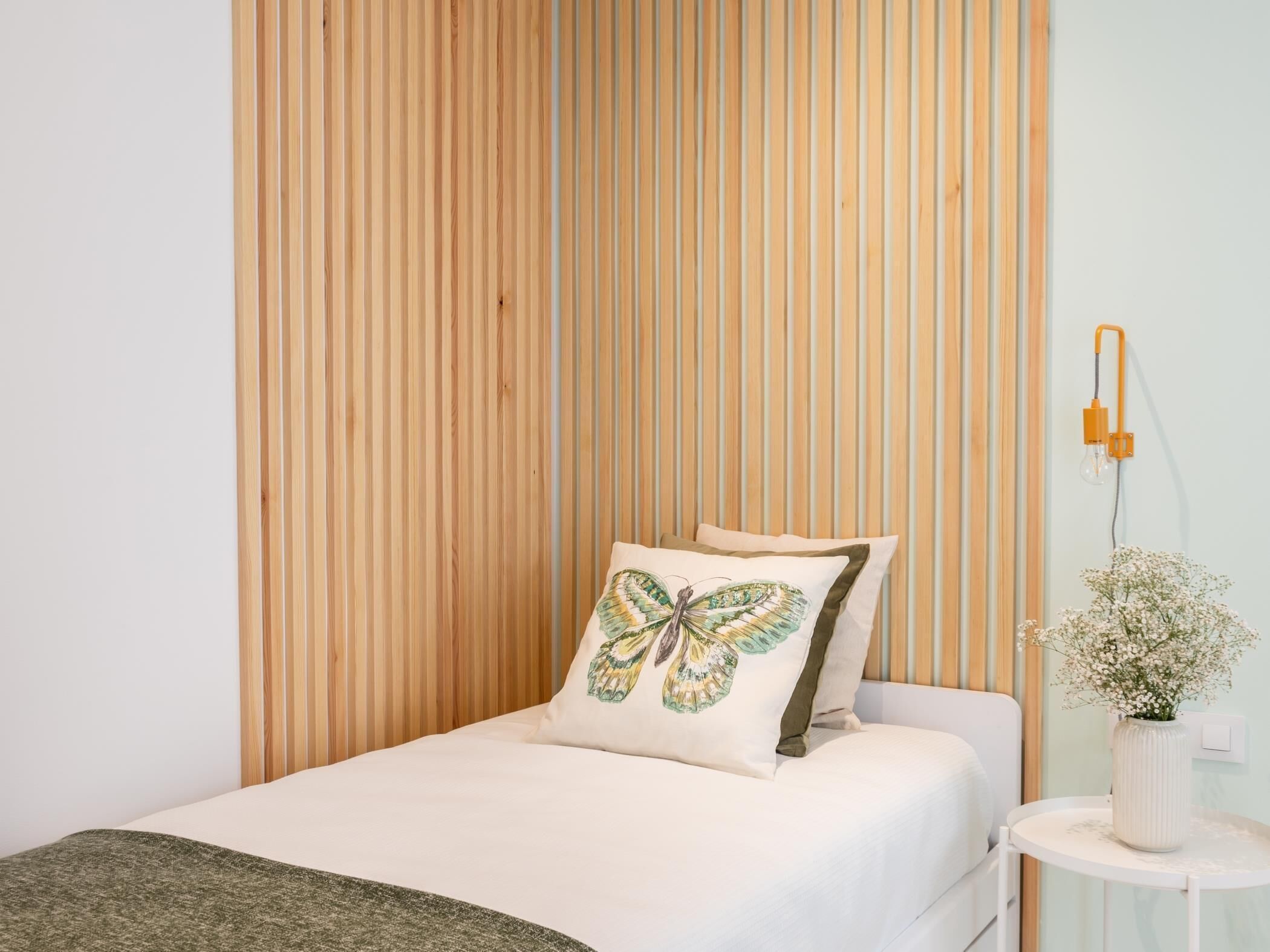 A bedroom with a bed, a wooden slat wall, a bedside table with a vase of flowers, and a wall-mounted lamp.