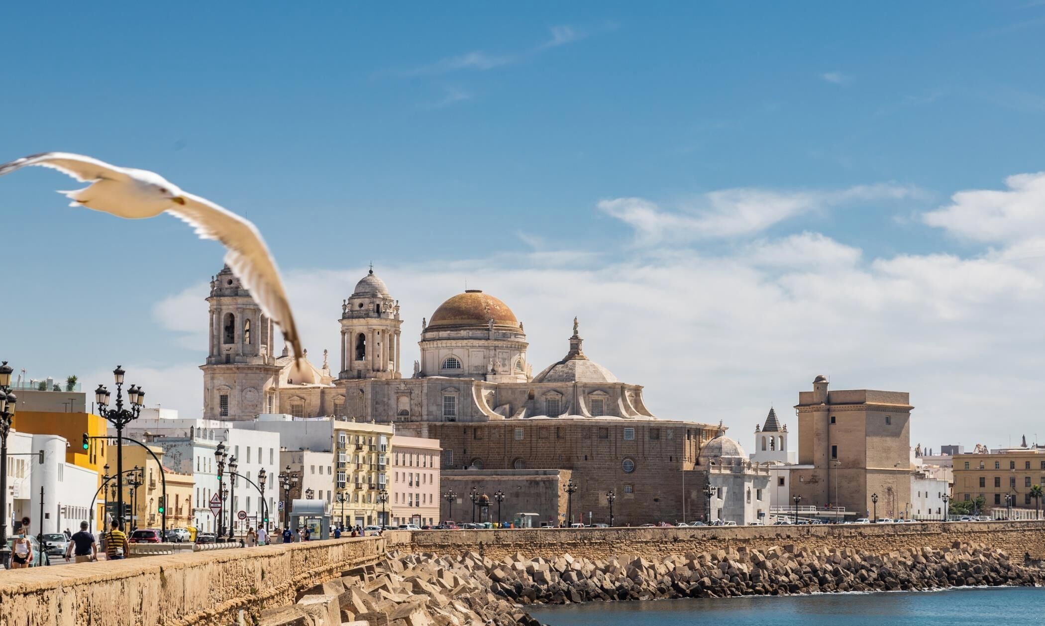 A coastal cityscape with historic architecture, including a prominent church with domes and bell towers, a rocky breakwater, and a clear blue sky.