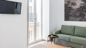 Un soggiorno con un divano verde, un piccolo tavolino con una pianta, una TV a parete e un balcone con ringhiera.