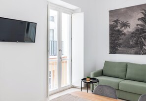 Ein Wohnzimmer mit einem grünen Sofa, einem kleinen schwarzen Tisch mit einer Pflanze, einem an der Wand befestigten Fernseher und einem Balkon mit Geländer.