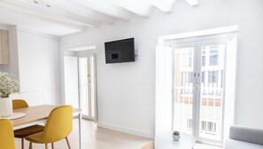 A modern kitchen with white cabinets, a dining table with yellow chairs, a flat-screen TV mounted on the wall, and a balcony with a sliding door. - Apartamento Villalobos 4 by Cádiz Time Apartments (Cádiz)
