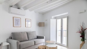 Een moderne woonkamer met een grijze bank, twee ingelijste foto’s, een vloerlamp, een klein houten tafeltje en een openslaande deur naar een balkon.