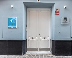 Ein Gebäudeeingang mit einer Doppeltür, einem blauen Schild mit der Aufschrift 'AT' und 'PLAYA' sowie einem 'Direct Alarma'-Schild.