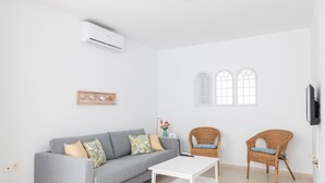 Un salon moderne avec un canapé gris, deux fauteuils en osier, une table basse blanche, un téléviseur fixé au mur et un climatiseur mural.