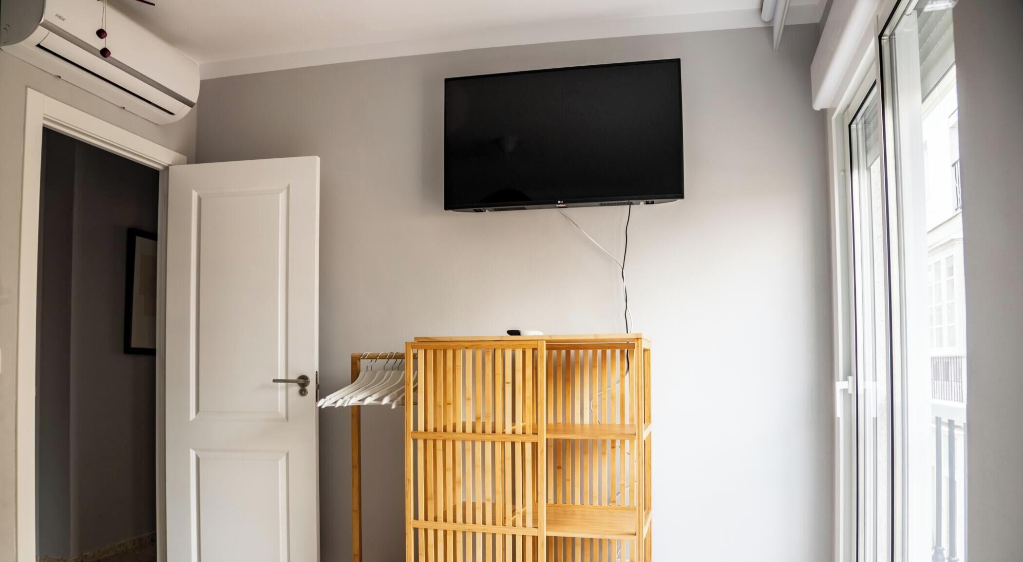A room with a white door, a wooden wardrobe, a mounted TV, and a window with blinds.