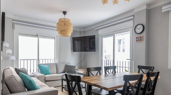 En moderne stue med sofa, spisebord, stole, fjernsyn og store vinduer.