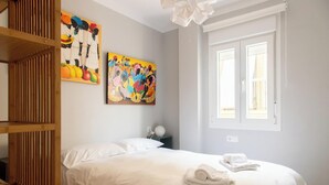Quarto com uma cama, duas pinturas, uma janela e uma estante de madeira.