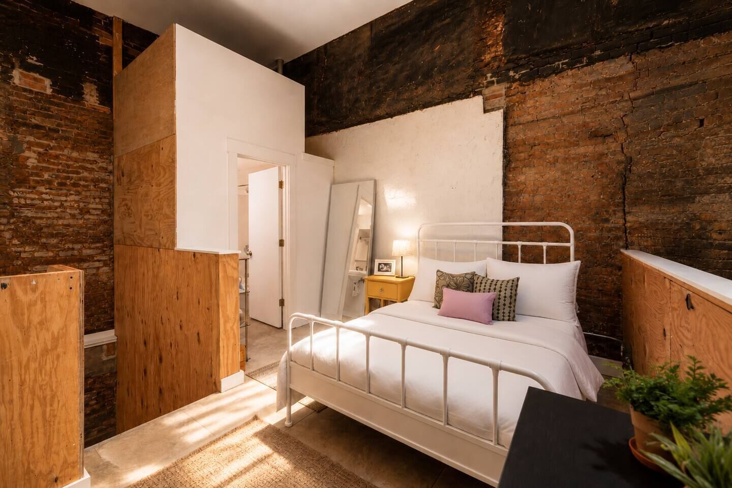 Un dormitorio con una cama de metal blanco, un armario de madera, una pared de ladrillo y una puerta blanca.
