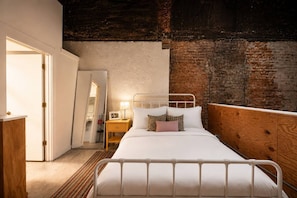 A bedroom with a white metal bed, a wooden headboard, a nightstand with a lamp, and a brick wall. - Artist Loft, Steps from TQL Stadium, Walk to All (Cincinnati)
