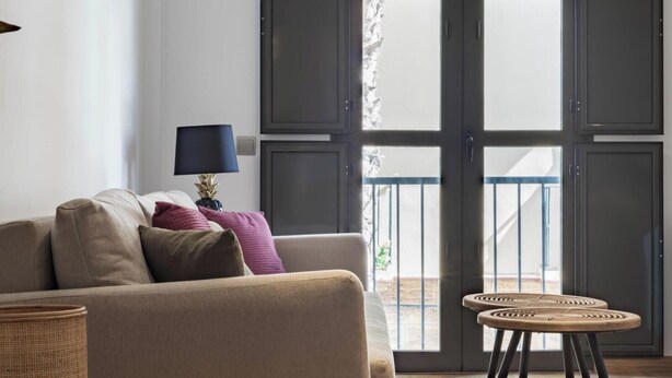 En stue med sofa, puder, lampe, et lille rundt bord og en skydedør, der fører ud til en balkon.