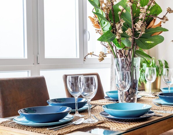 Ein Esstisch ist gedeckt mit blauen Tellern, klaren Weingläsern und einer Vase mit grünen Blättern und weißen Blumen.