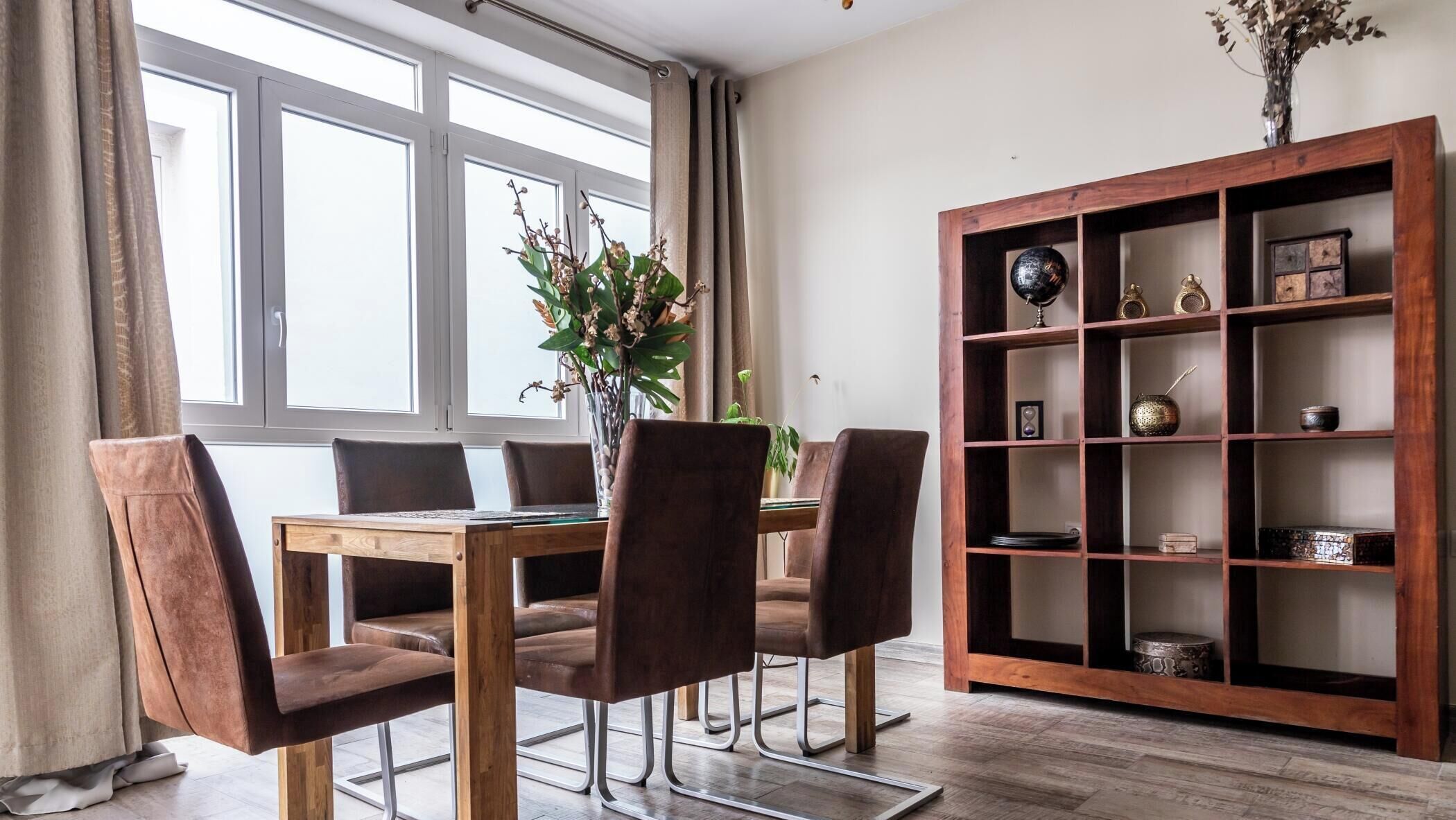 Un comedor con una mesa y sillas de madera, un jarrón con flores y una estantería de madera con adornos.