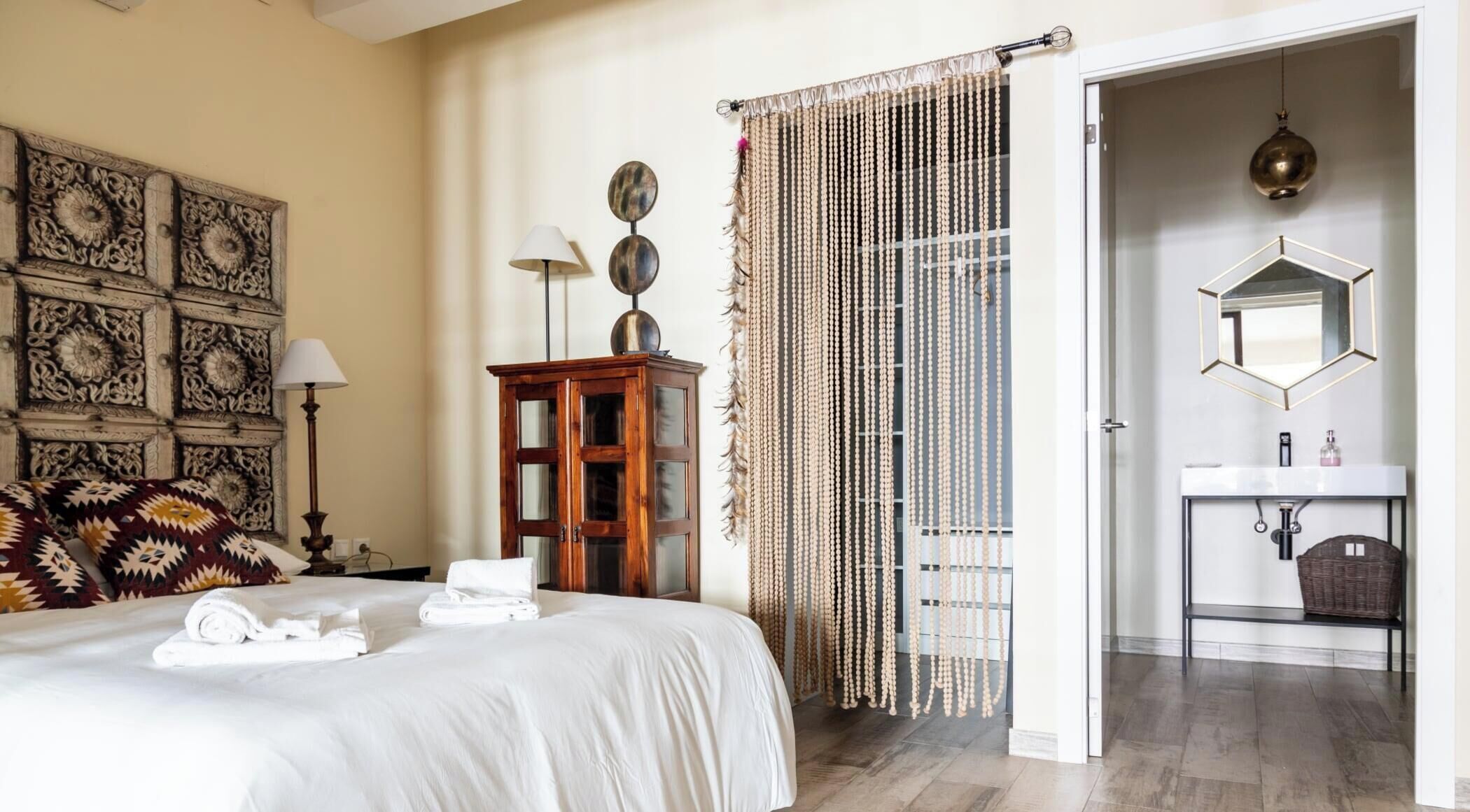 Une chambre d’hôtel comprenant un lit, une armoire en bois, un mur décoratif, un rideau en perles et une salle de bain avec un miroir et un lavabo.