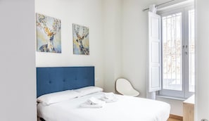 A bedroom with a bed, white pillows, a blue headboard, two wall-mounted paintings, a white bedside table, and a window with a view. - Nature Home by Cadiz Time (Cádiz)