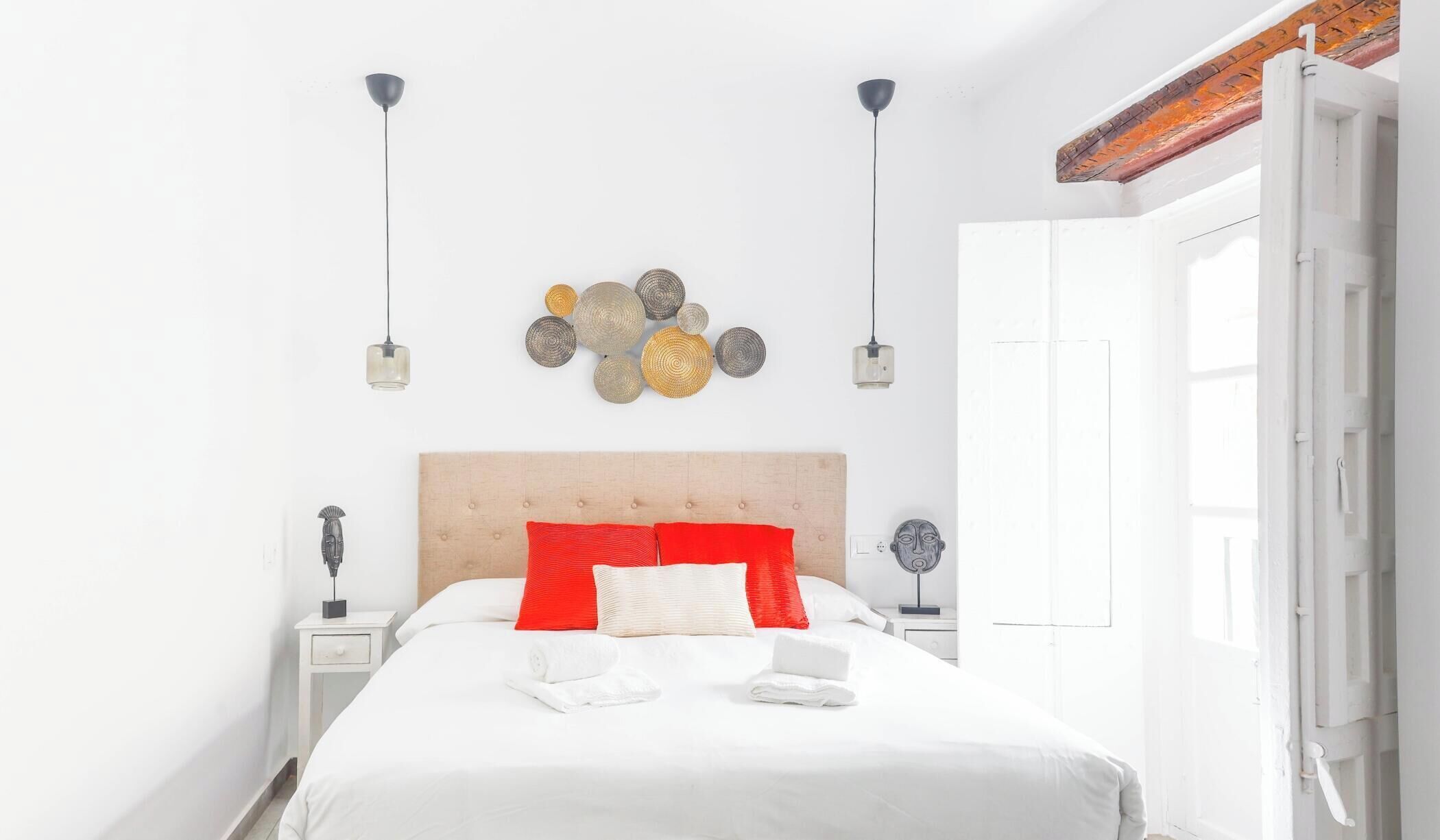 A neatly made bed with white bedding and red pillows, a wooden headboard, and a wall-mounted decorative item.