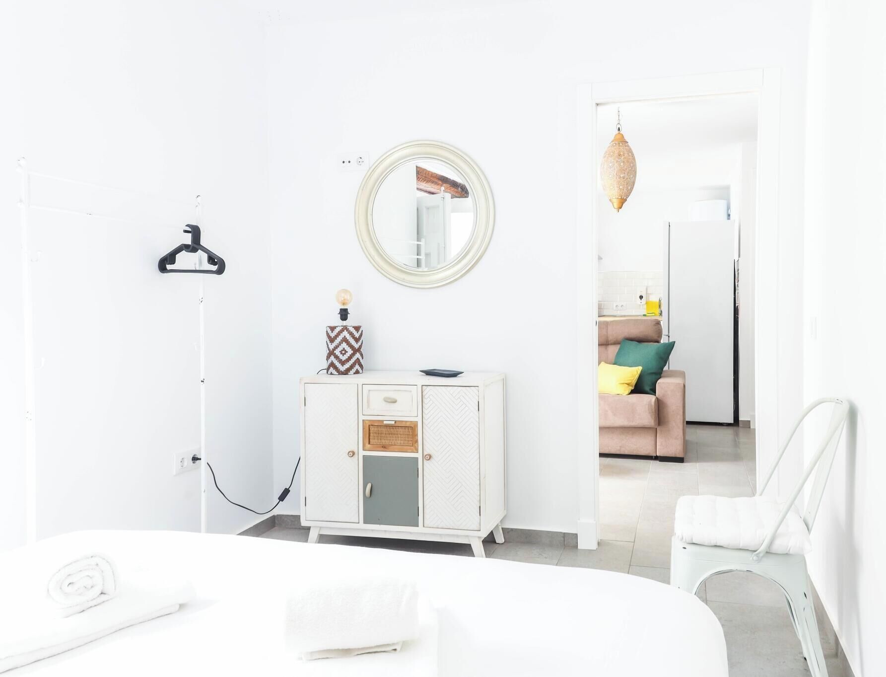 A modern, minimalist bedroom with a bed, a white dresser, a hanging light, and a mirror.