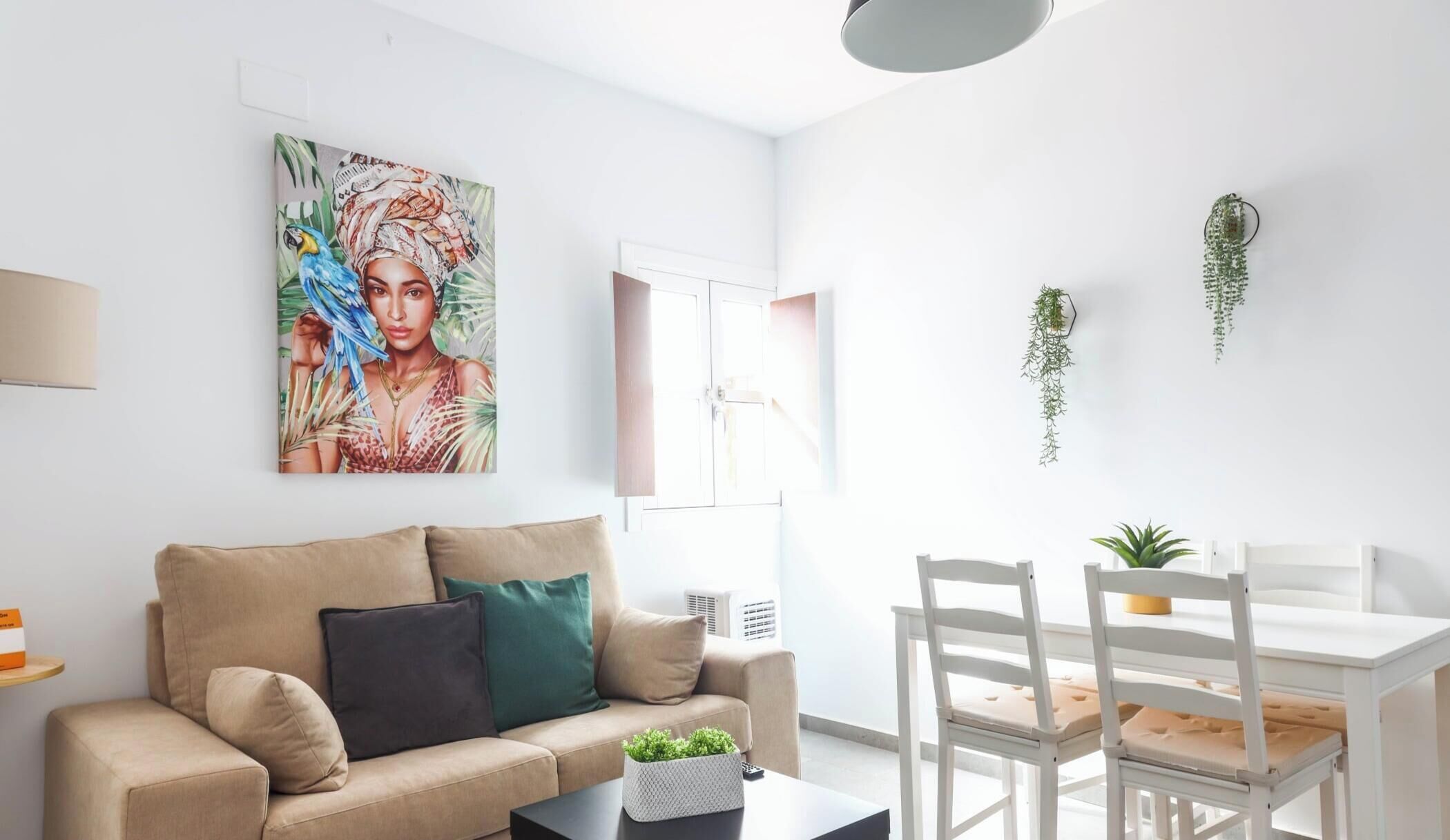 En stue med en beige sofa, et sofabord, et hvidt spisebord med stole, et billede på væggen og et vindue med skodder.
