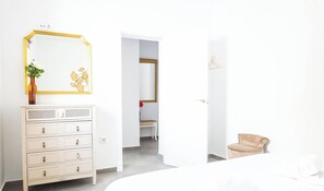 Ein Zimmer mit einem weißen Schminktisch, einem Spiegel, einer Vase mit Pflanze, einem beigen Sessel und einer Tür zu einem anderen Raum.