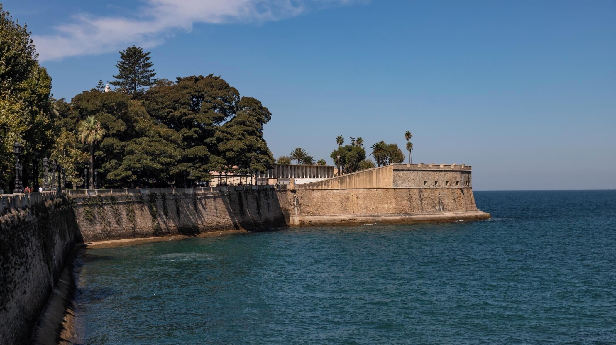 A coastal fortification with a stone wall, trees, and a clear sky.