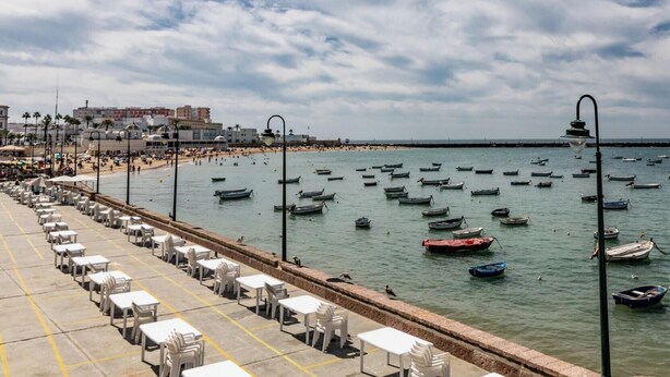 Paesaggio costiero con spiaggia, barche e una passeggiata dotata di tavoli e sedie.